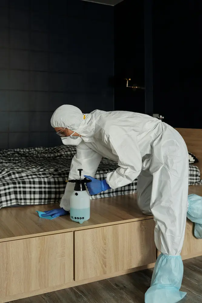 Specialist in white hazmat suit spraying disinfectant in bedroom during sanitization