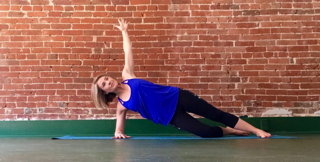 Heather Hausman in a blue tank top performs a side plank exercise on a turquoise yoga mat against a rustic brick wall.