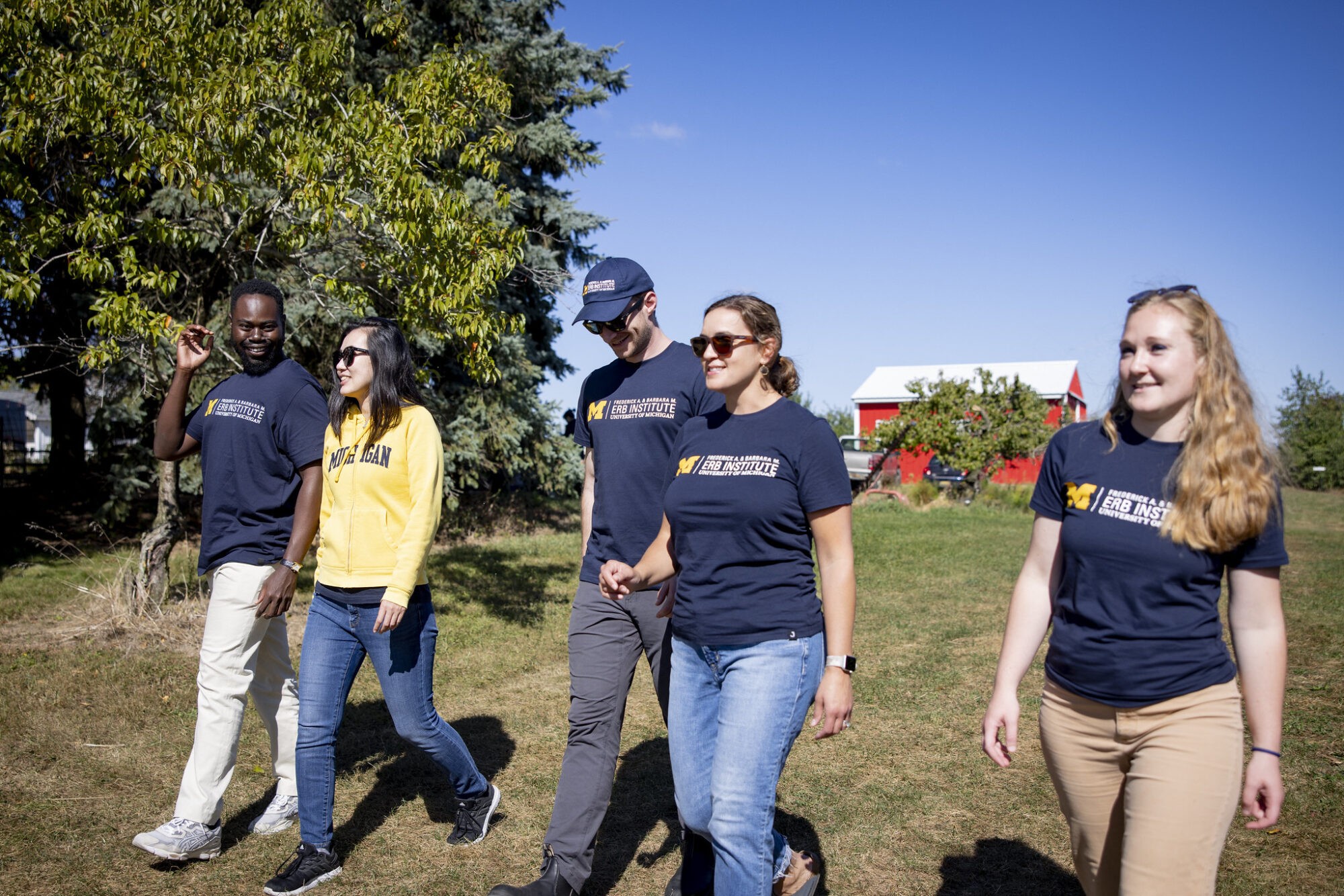 A group of five people walking together outdoors, wearing matching shirts, on a sunny day with trees in the background.