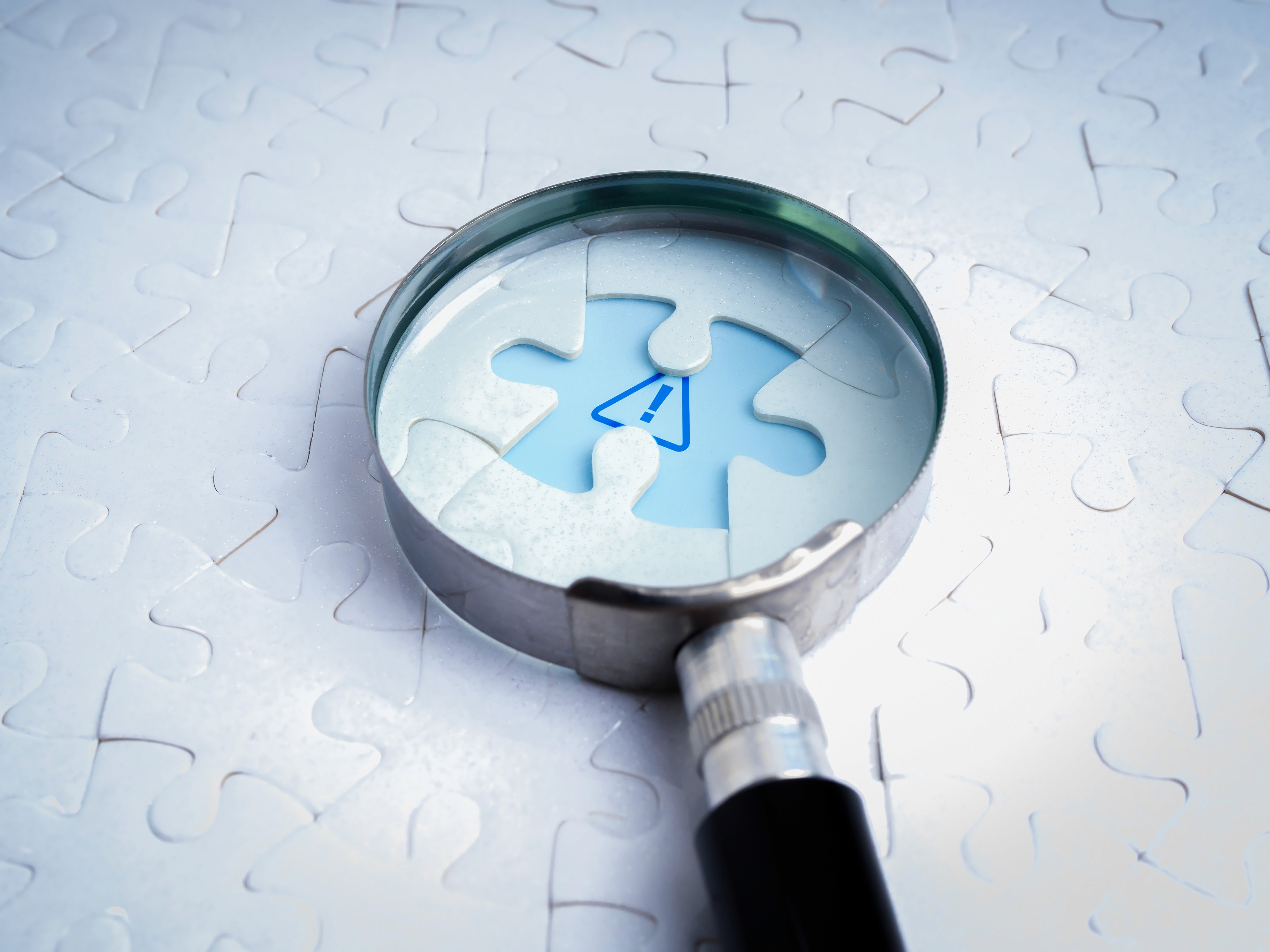 Business solutions, problems found, problem solving concept. Close-up blue caution sign, graphic on space of last missing piece, focus in magnifying glass lens, on white jigsaw puzzle background.