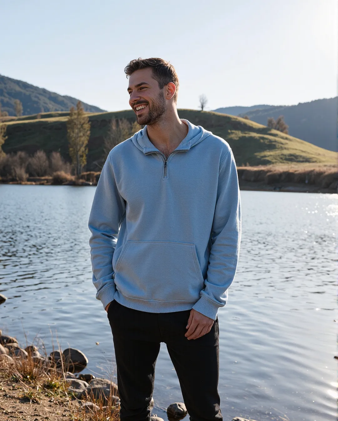 Person in light blue hoodie smiling by a lake with rolling hills in background, captured in warm natural sunlight