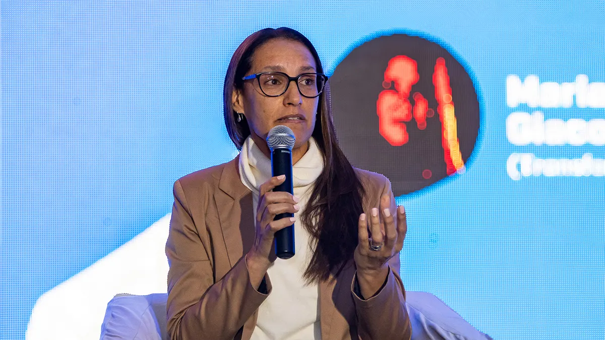 A woman wearing glasses and a brown blazer speaks into a microphone on stage.