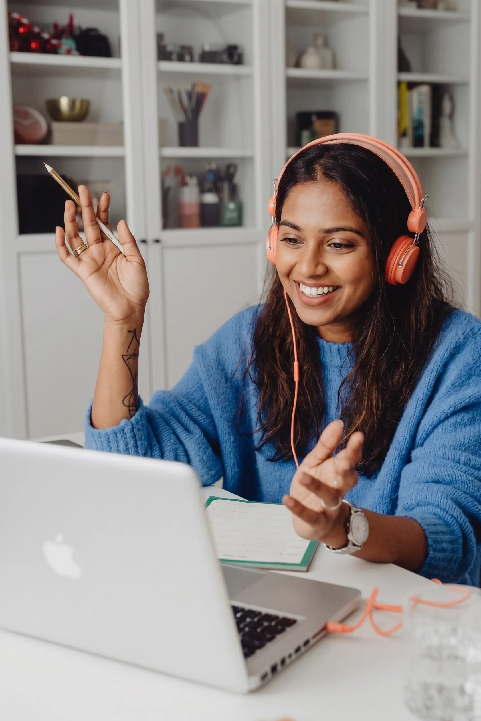 A smiling blonde female teacher wearing pink headphones and a blue sweater gestures with her hand while interacting during an online class on her laptop.
