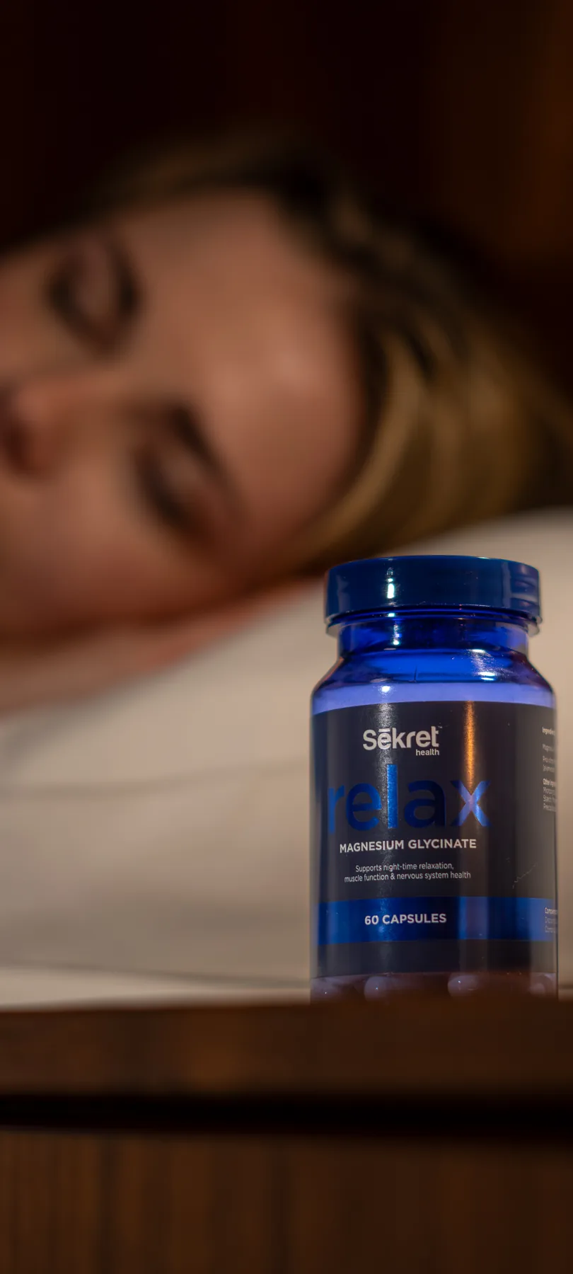 A bottle of Sēkret Relax capsules on a bedside table while a woman sleeps behind it