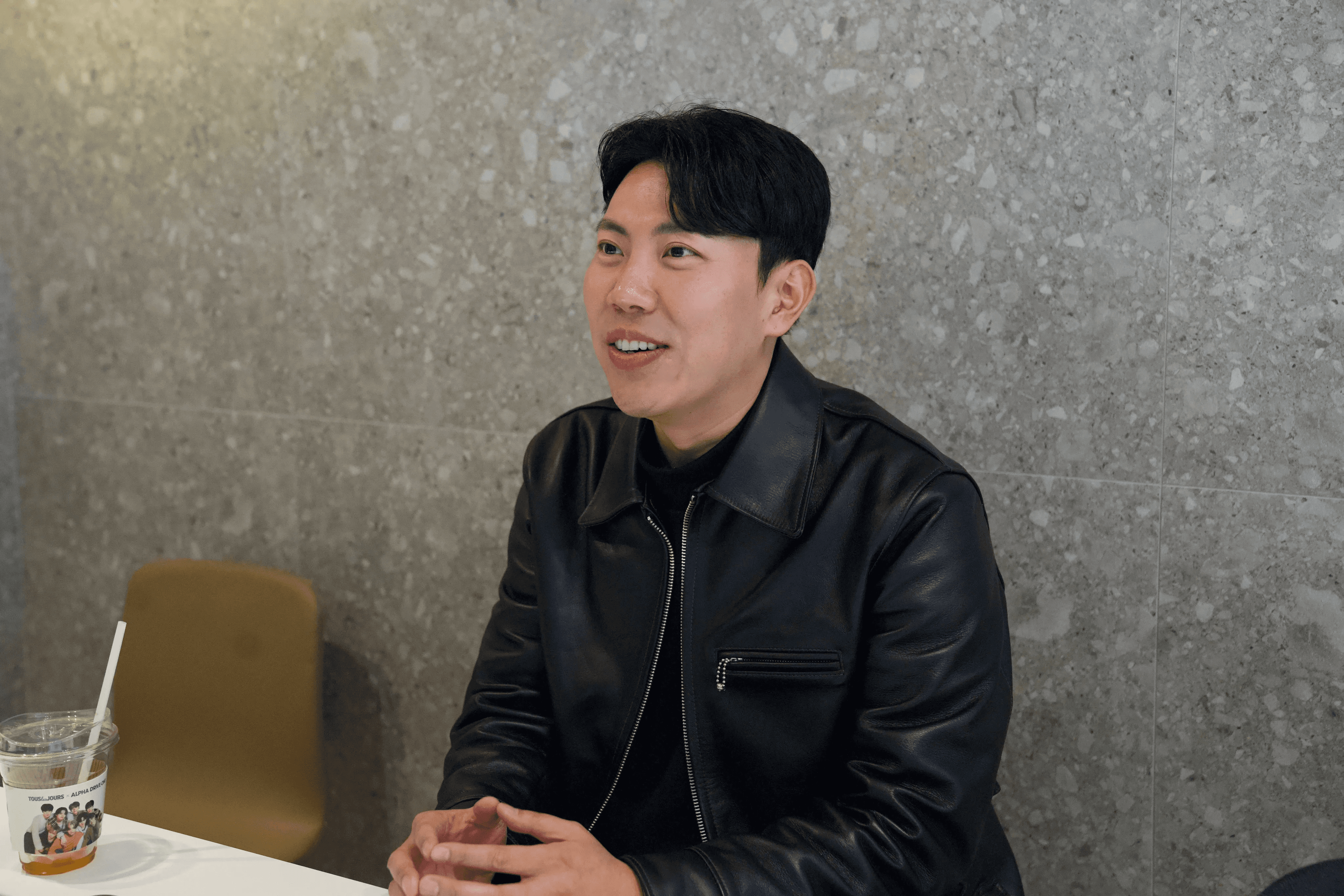 회색 테라조 벽을 배경으로 검정 가죽 재킷을 입은 남성이 테이블에 앉아 측면을 바라보며 밝게 웃고 있는 모습. 테이블 위에 음료컵이 놓여 있습니다.