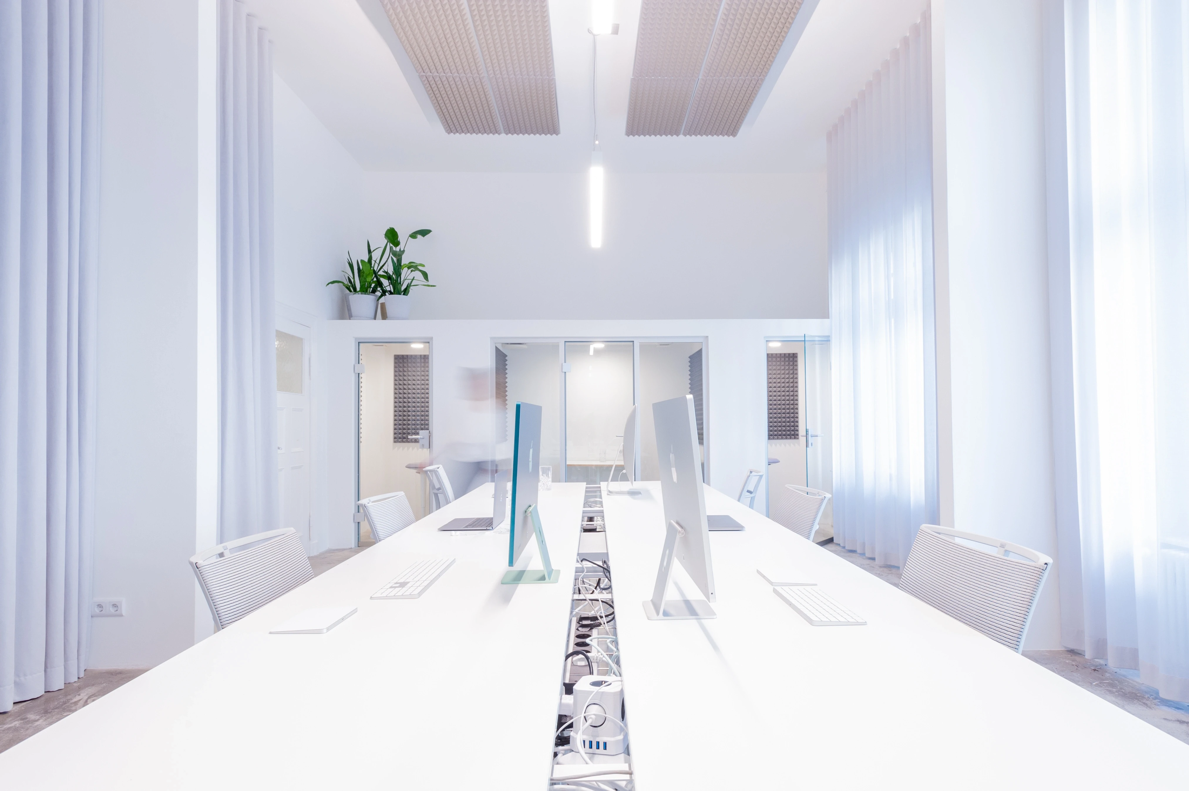Symmetrical view down a long white desk row at BATTLE ROYAL with white iMacs back to back, a patterned cable tray in the middle, sheer curtains and a tall white ceiling.