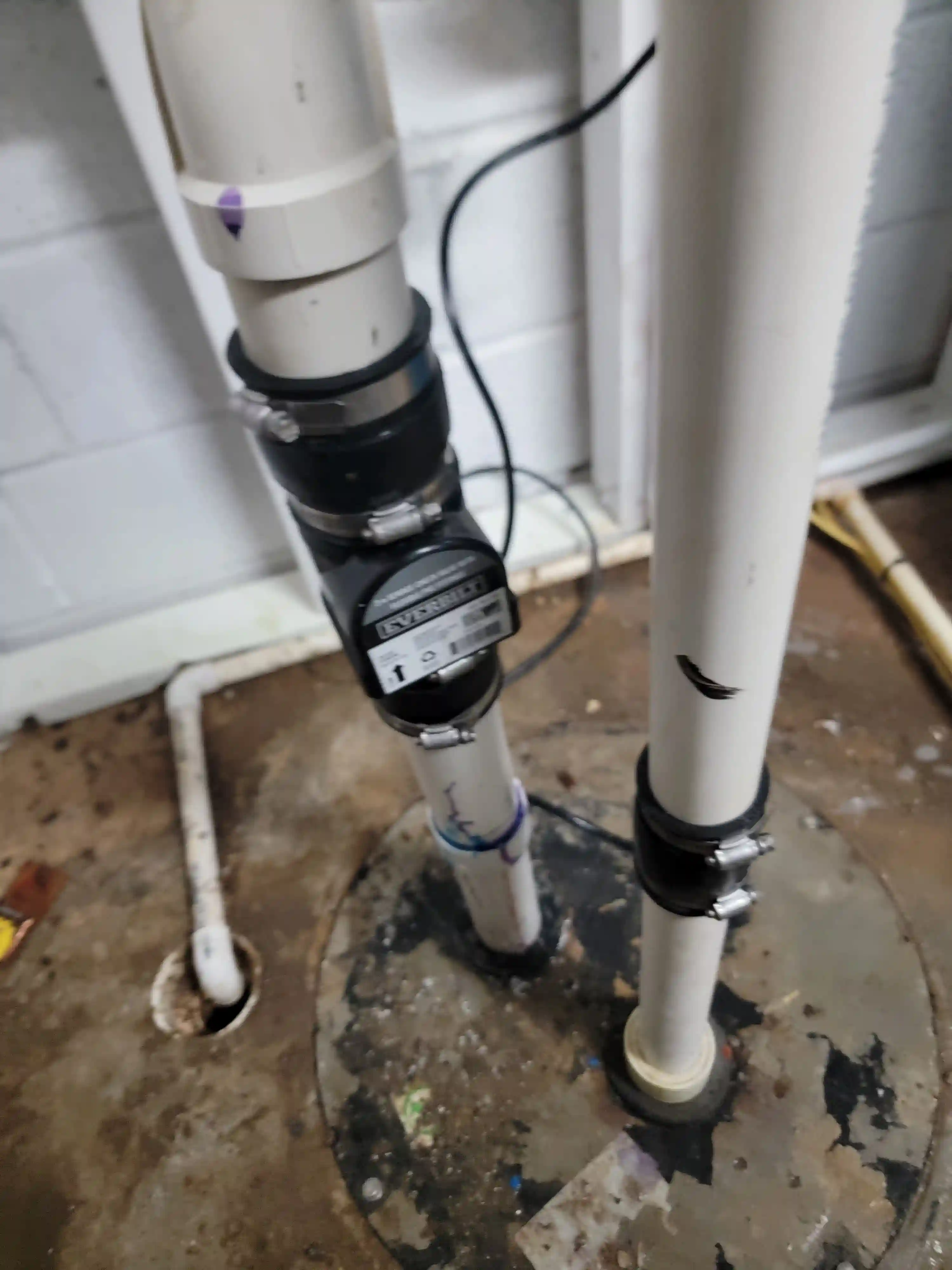 A close-up of a black check valve secured to PVC discharge pipes, inspected during a basement sump pump repair.