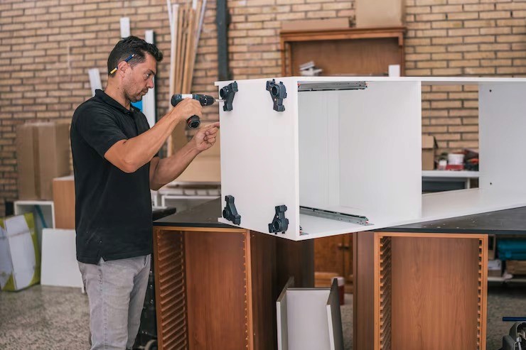 A carpenter wearing safety glasses screws parts of white cabinets together with a drill on a workbench.