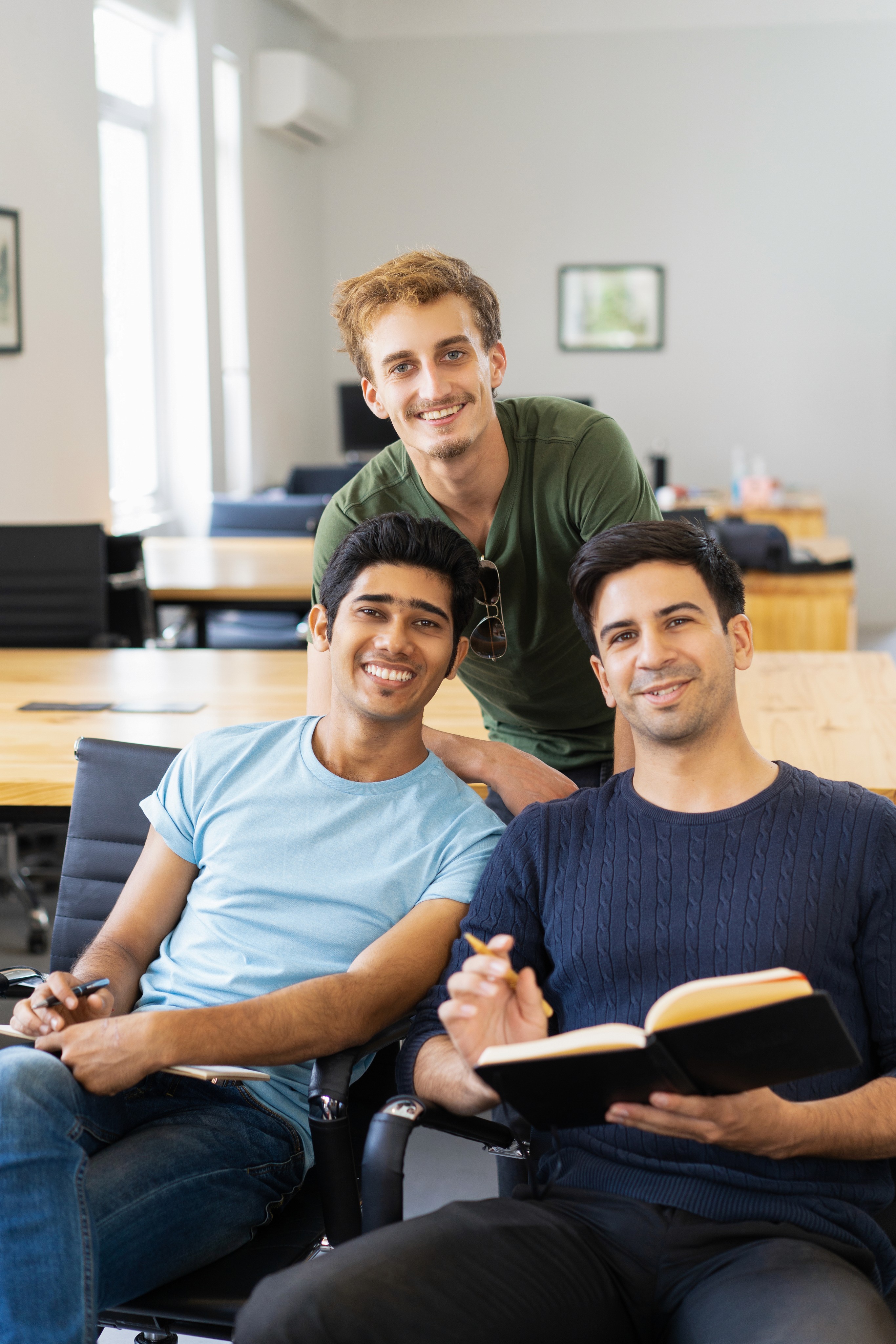 Three young men in a modern office, with one seated holding a tablet.