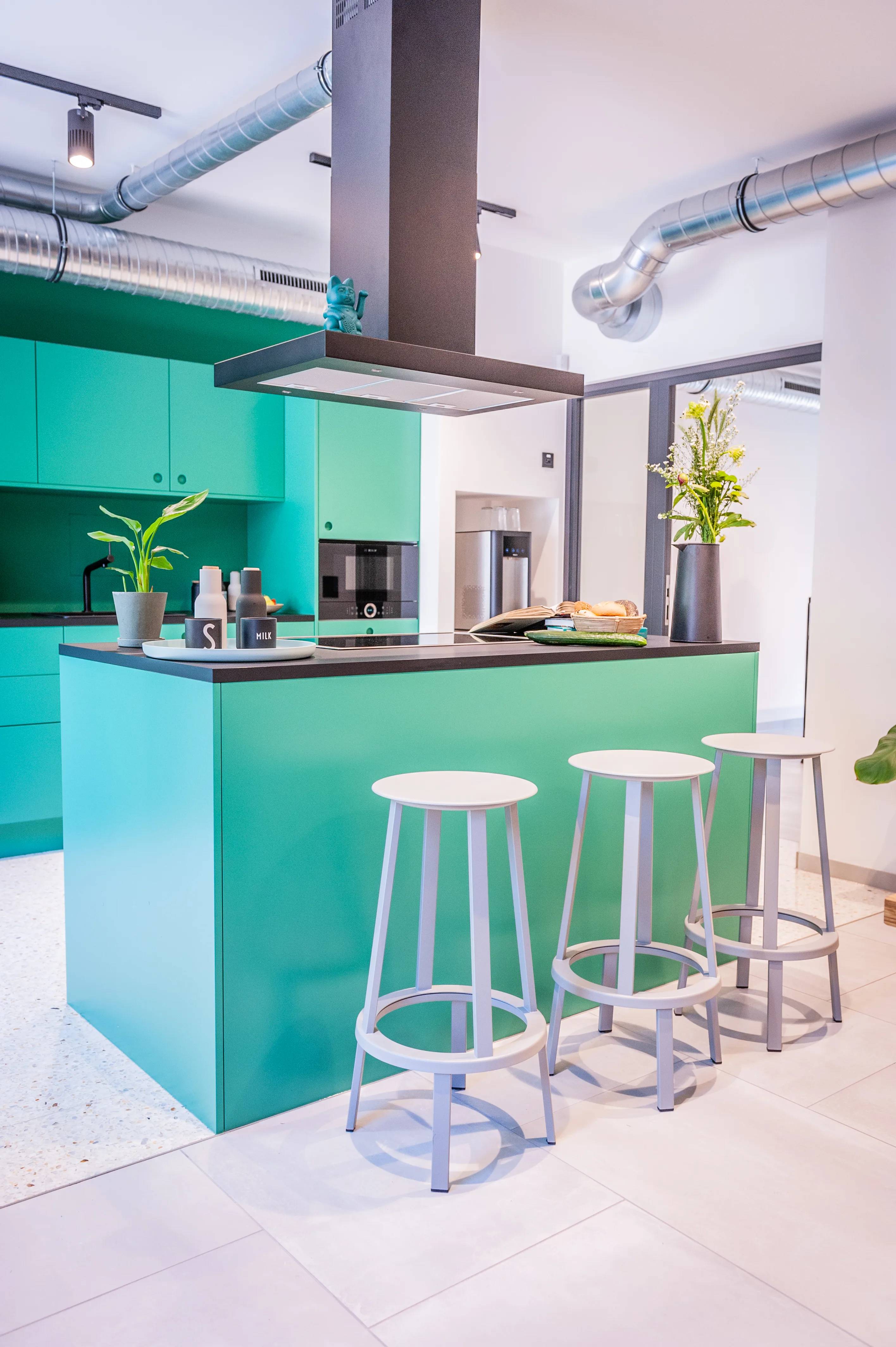 Mint-green kitchen island at EHEALTH-TEC with black countertop, industrial extractor hood, exposed ventilation ducts, three grey bar stools and terrazzo flooring.