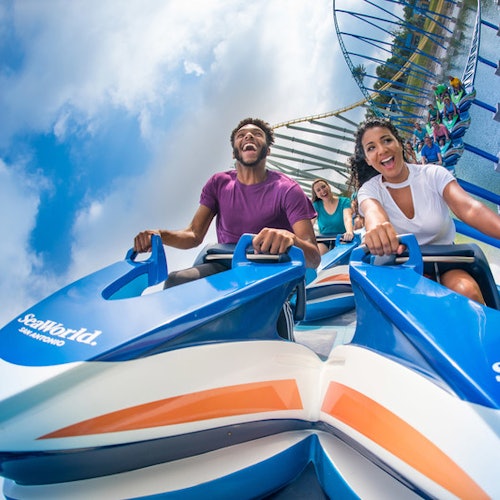 Two people smiling and holding on while riding a blue and white rollercoaster at SeaWorld on a sunny day.