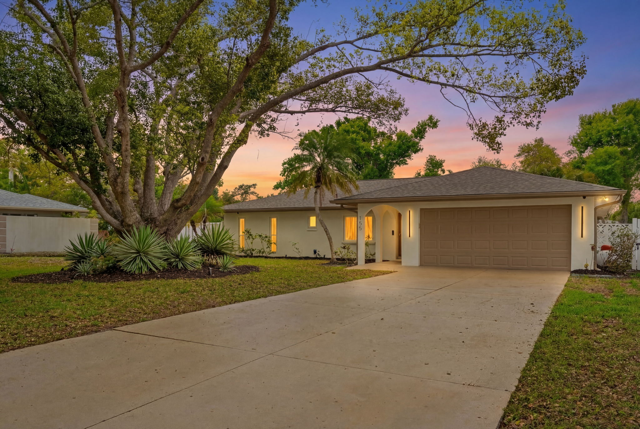 Single-story stucco home near Sarasota at dusk with warm lit windows, a two-car garage, a large spreading tree and a wide driveway under a pink and blue sky