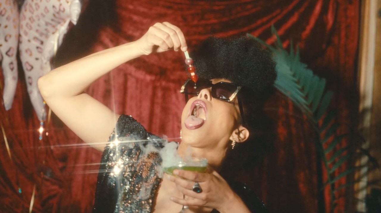 Woman with sunglasses licking a dropper over a smoking green drink against a red curtain.