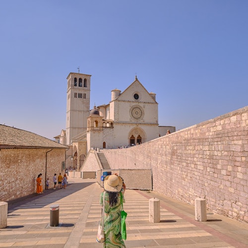 A person in a green outfit and hat walks toward a historic church with a bell tower under a clear blue sky.