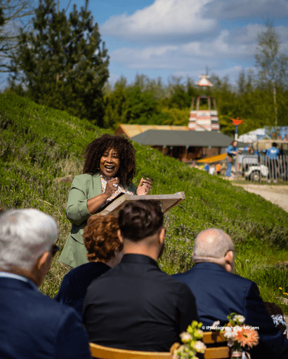 Een zelfstandig trouwambtenaar spreekt met enthousiasme tijdens een huwelijksceremonie in de buitenlucht, terwijl gasten aandachtig luisteren op een zonnige trouwdag.
