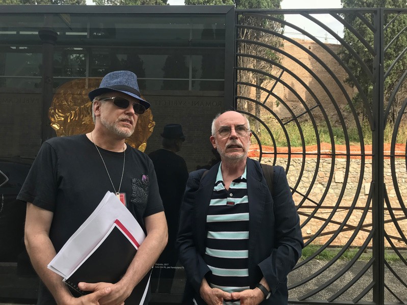 A photo of two men sitting in front of a gate with some round grills on it.