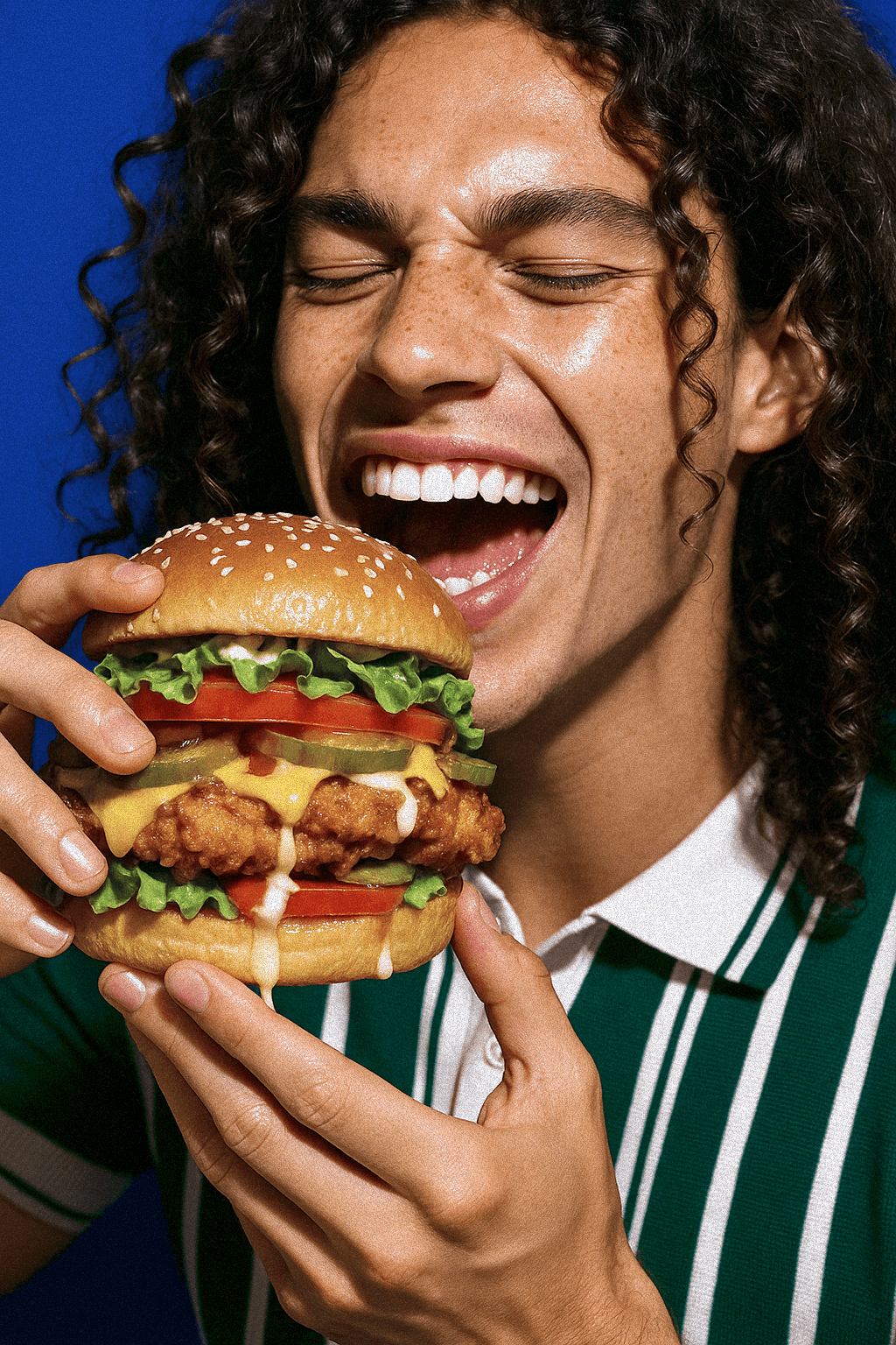 Smiling person holding a large burger with fried chicken, cheese, lettuce, tomato, pickles, and sauce, about to take a bite.