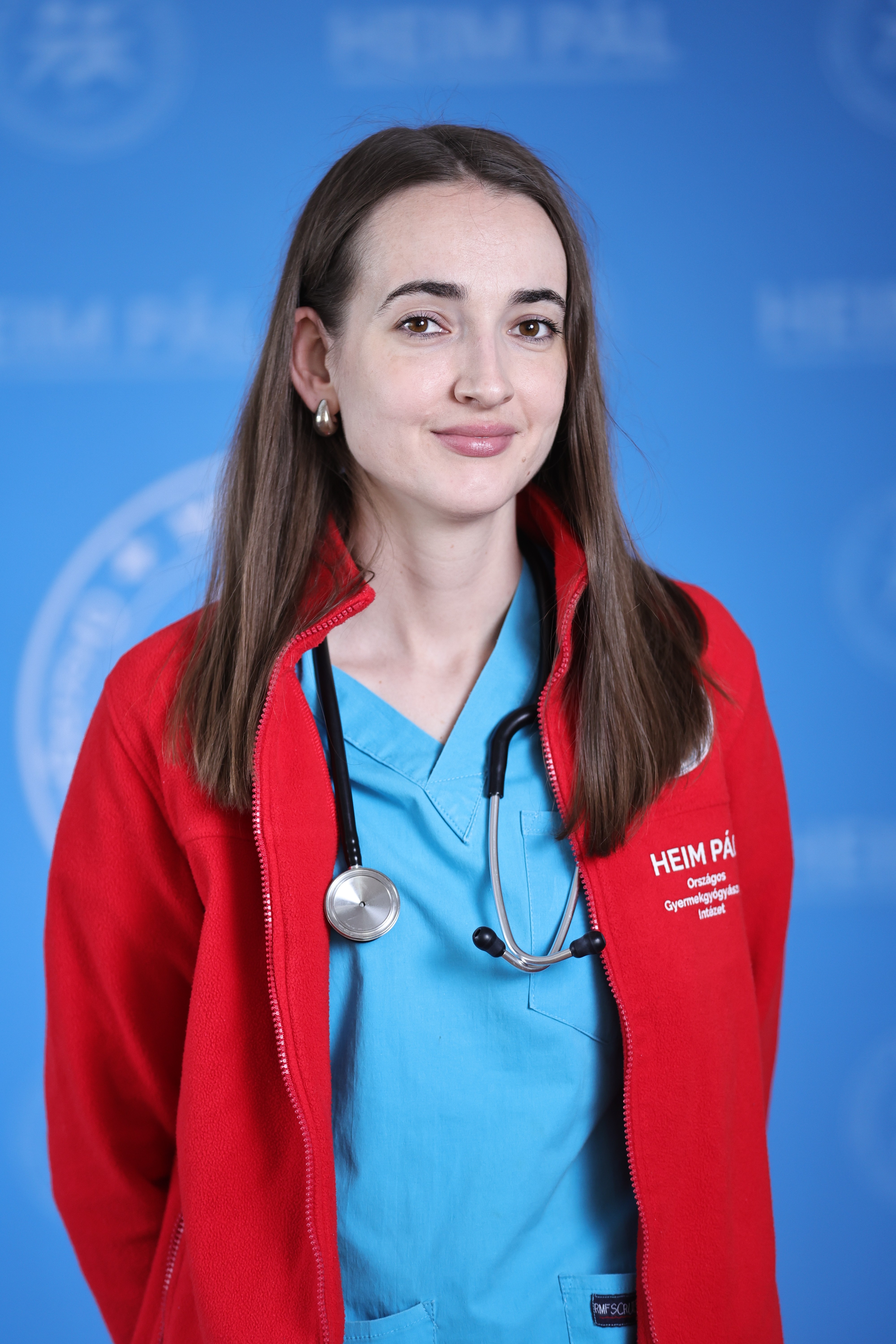 Barna hajú nő piroskék orvosi ruhában sztetoszkóppal kék háttér előtt.