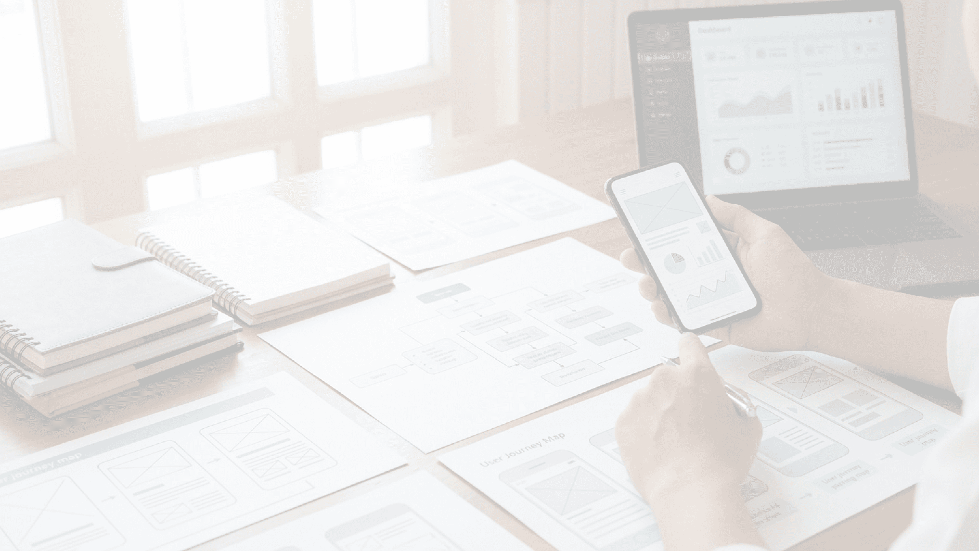 Fotografía de primer plano de un usuario sosteniendo un teléfono con un prototipo de baja fidelidad, sobre un escritorio con wireframes en papel y una laptop que muestra el mismo diseño digitalizado.