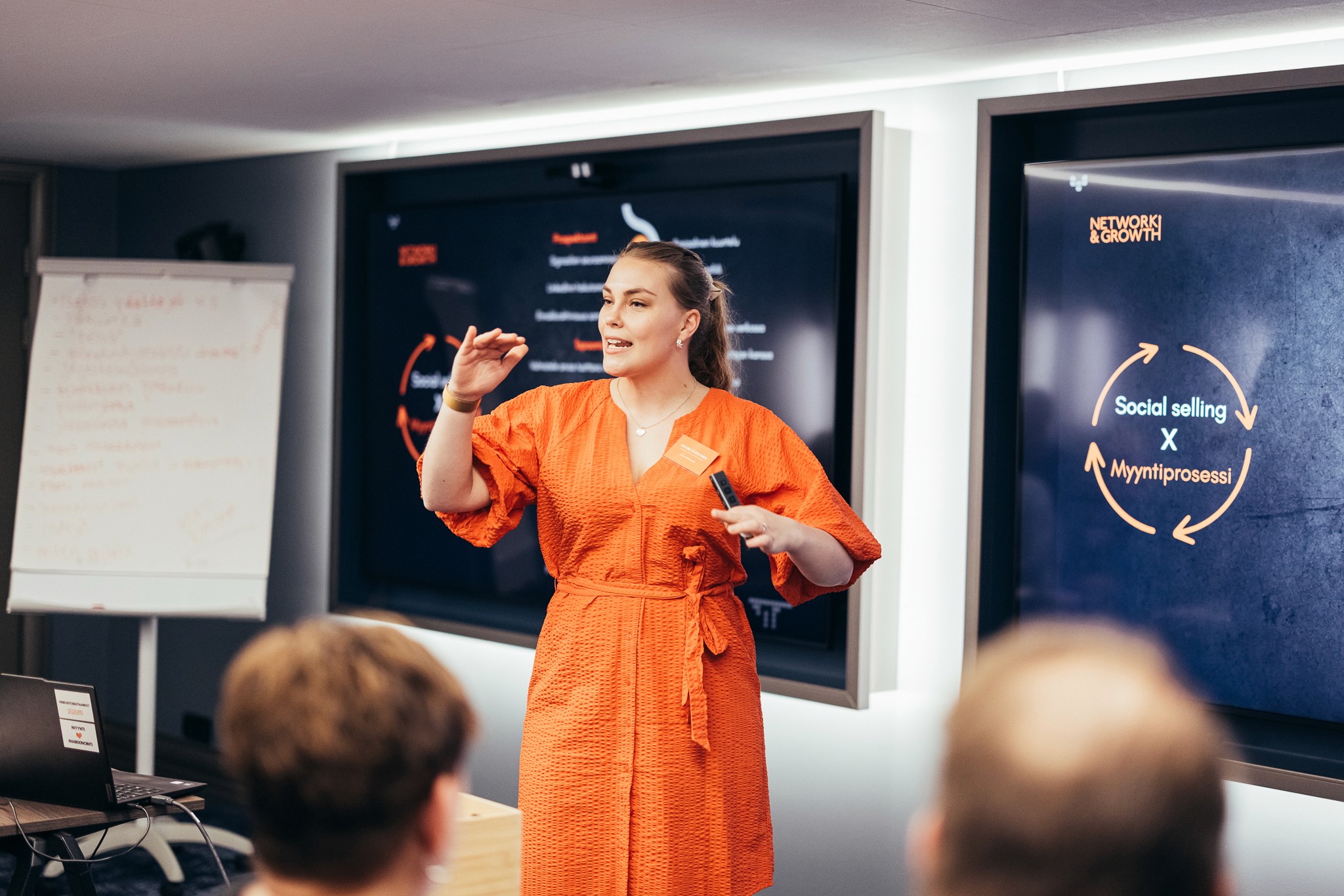 Oranssissa mekossa esiintyjä esittelee konferenssissa; kahdella näytöllä teksti “Social selling × Myyntiprosessi”; yleisö etualalla.