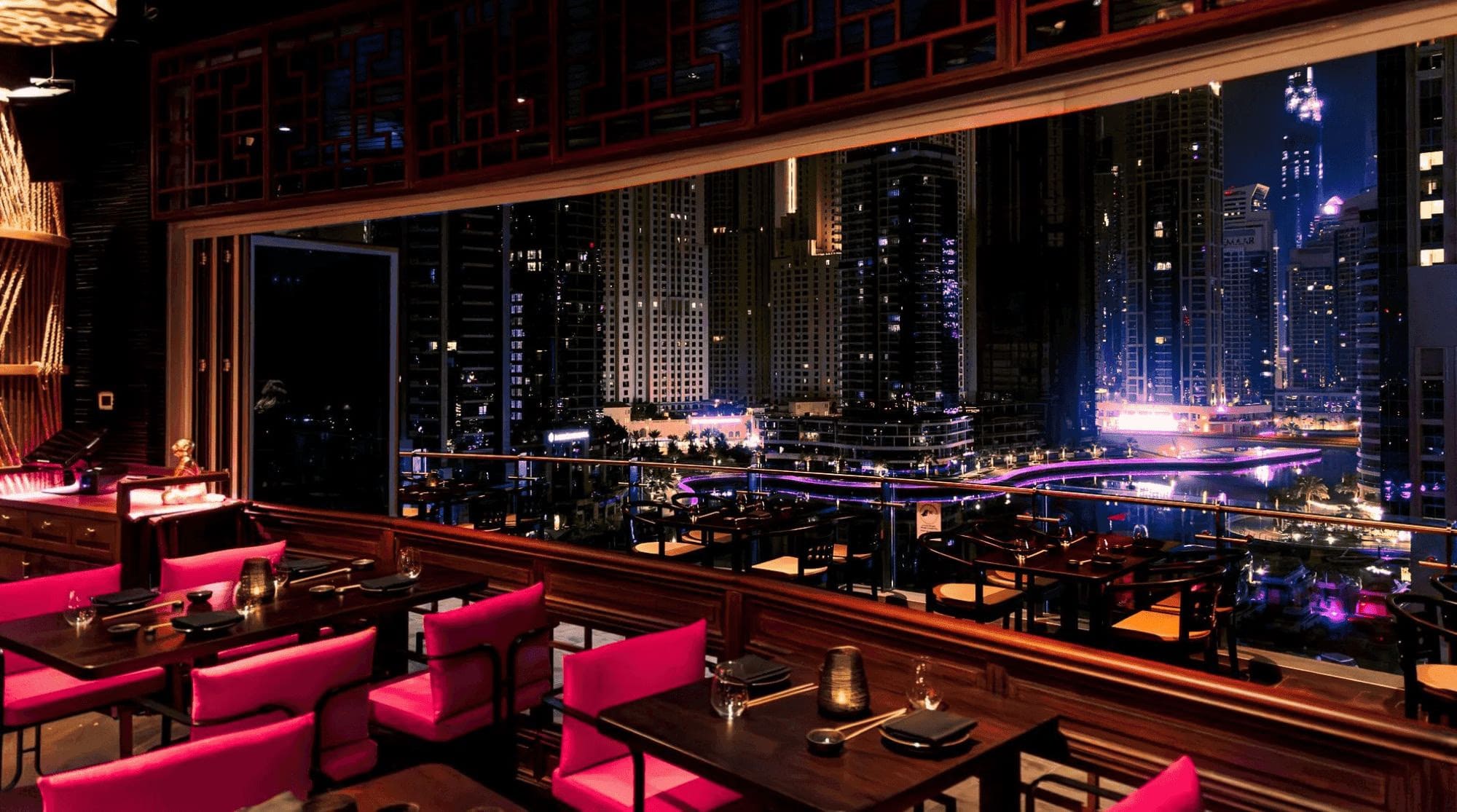 A restaurant in Dubai Marina with indoor seating and a large open window overlooking the illuminated marina skyline at night.