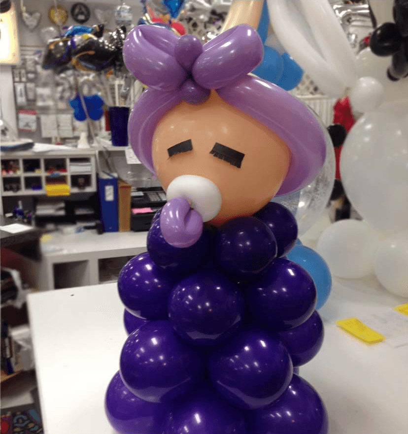 A creative balloon sculpture resembling a baby, with a purple balloon body and matching pacifier, is displayed on a store counter filled with various party supplies and decorations.