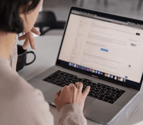 책상 앞에서 맥북 노트북을 사용 중인 사람의 뒷모습입니다. 화면에 웹사이트 혹은 문서를 띄워두고 한 손으로는 터치패드를 조작하며 내용을 검토하는 모습입니다.