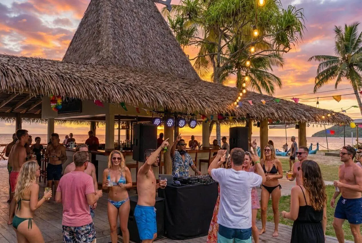 Guests dancing at a lively thatched-roof beachfront bar party with a DJ at sunset, Uprising Beach Resort Fiji
