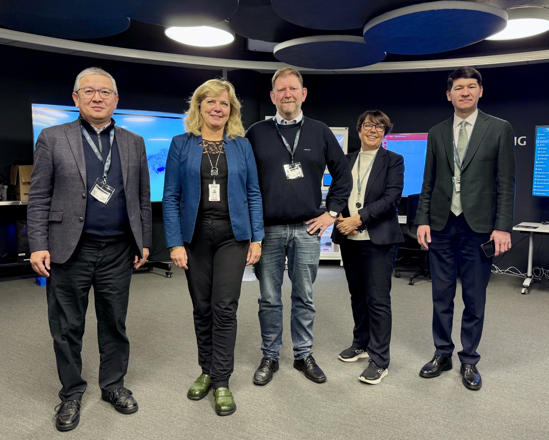 A group of five individuals standing together in a modern office setting with screens in the background.