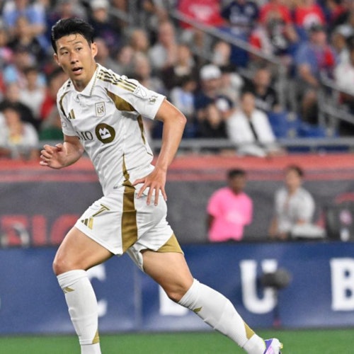 A soccer player in a white and gold uniform runs on the field during a game. Spectators are visible in the background.
