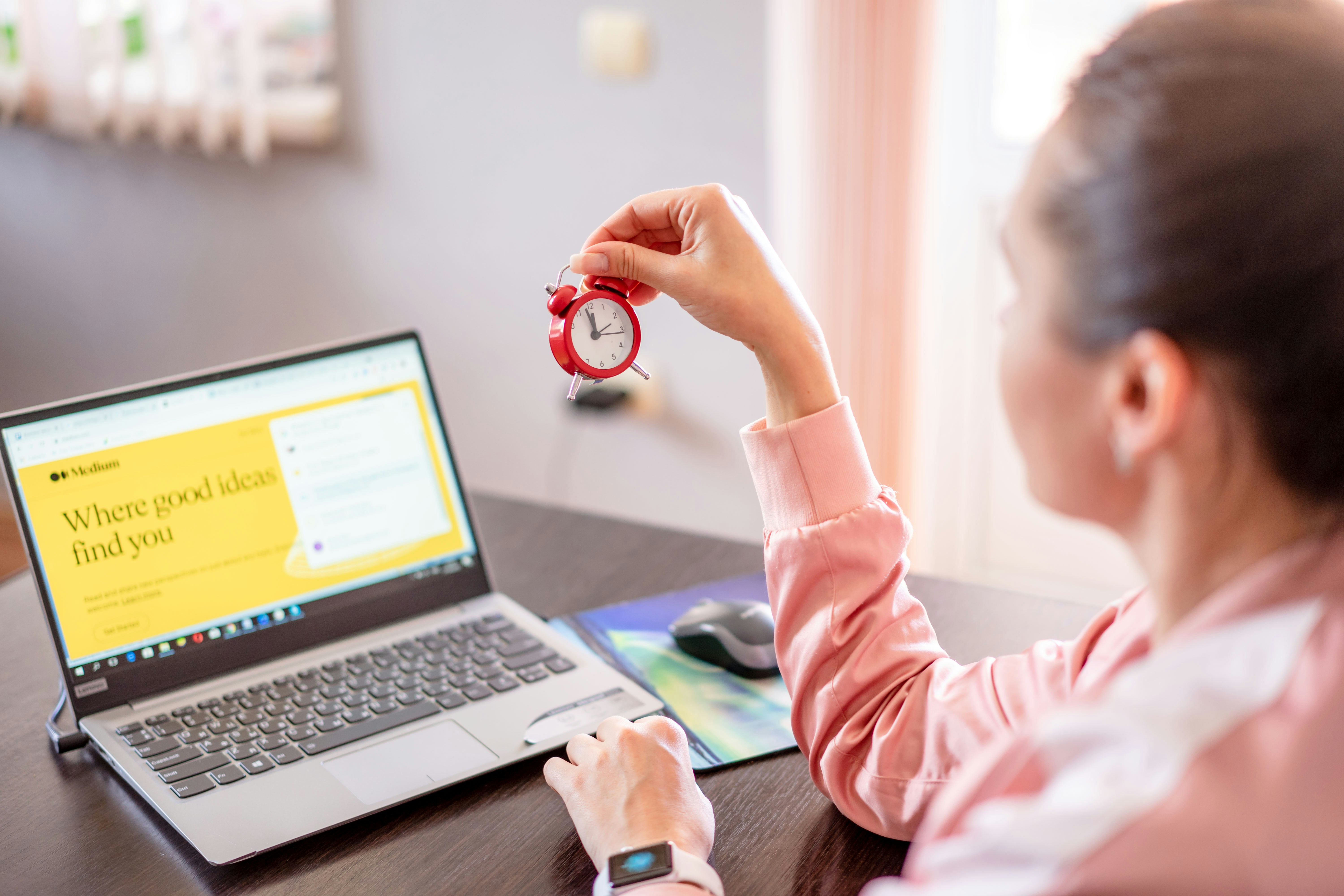 A woman looks at an open laptop. In one hand she holds an alarm clock.