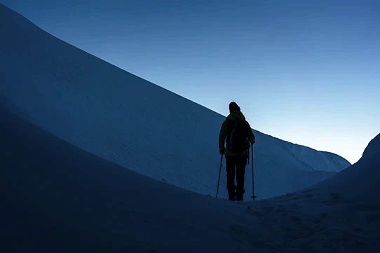 Sylwetka turysty z kijkami trekkingowymi na śnieżnym szlaku w górach o zmierzchu, z jasnym niebem w tle