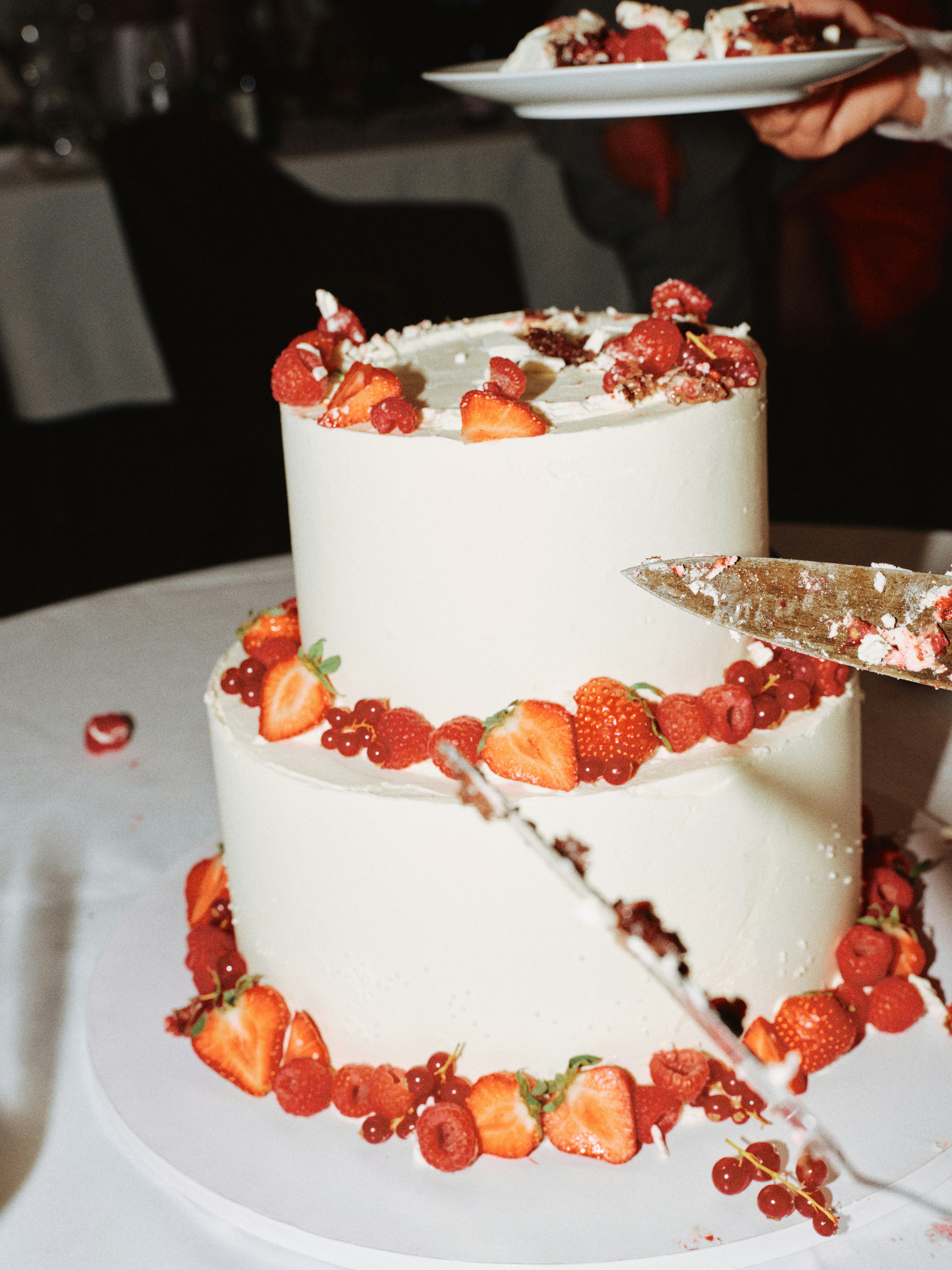 Dramatic close-up of a knife slicing through a two-tier white wedding cake decorated with strawberries and red currants, a plated slice visible in the background
