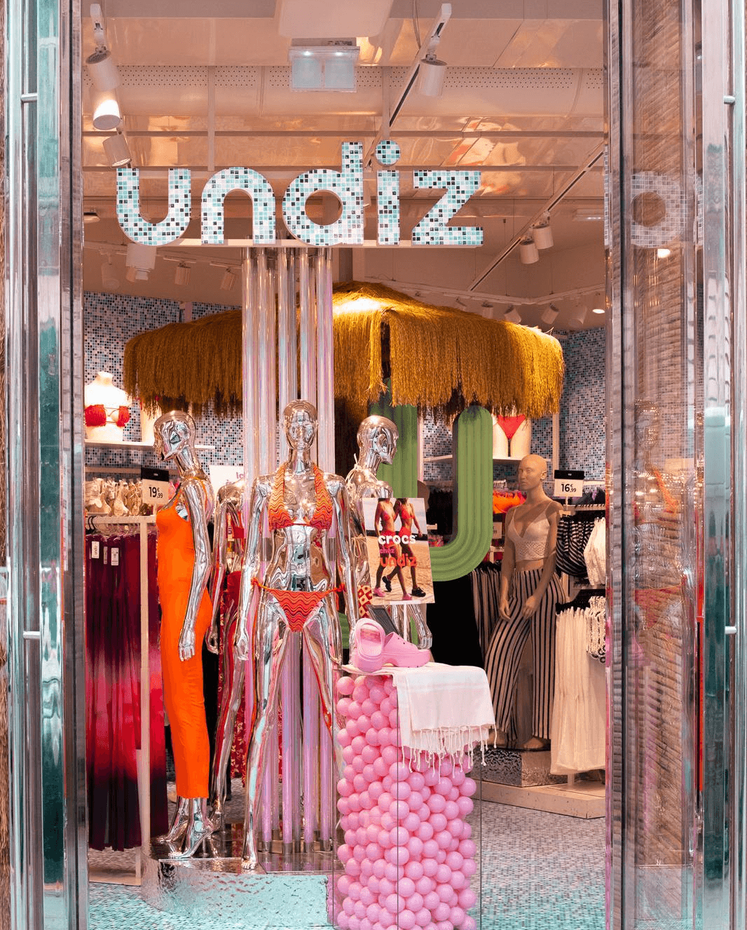 Entrée du magasin Undiz décorée pour l'été 2024 avec mannequins, boules roses et parasols, enseigne lumineuse visible.