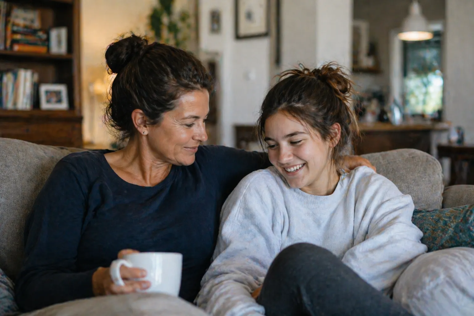 A woman holding a white mug smiles down at a smiling teen girl sitting beside her on a sofa in a cozy home