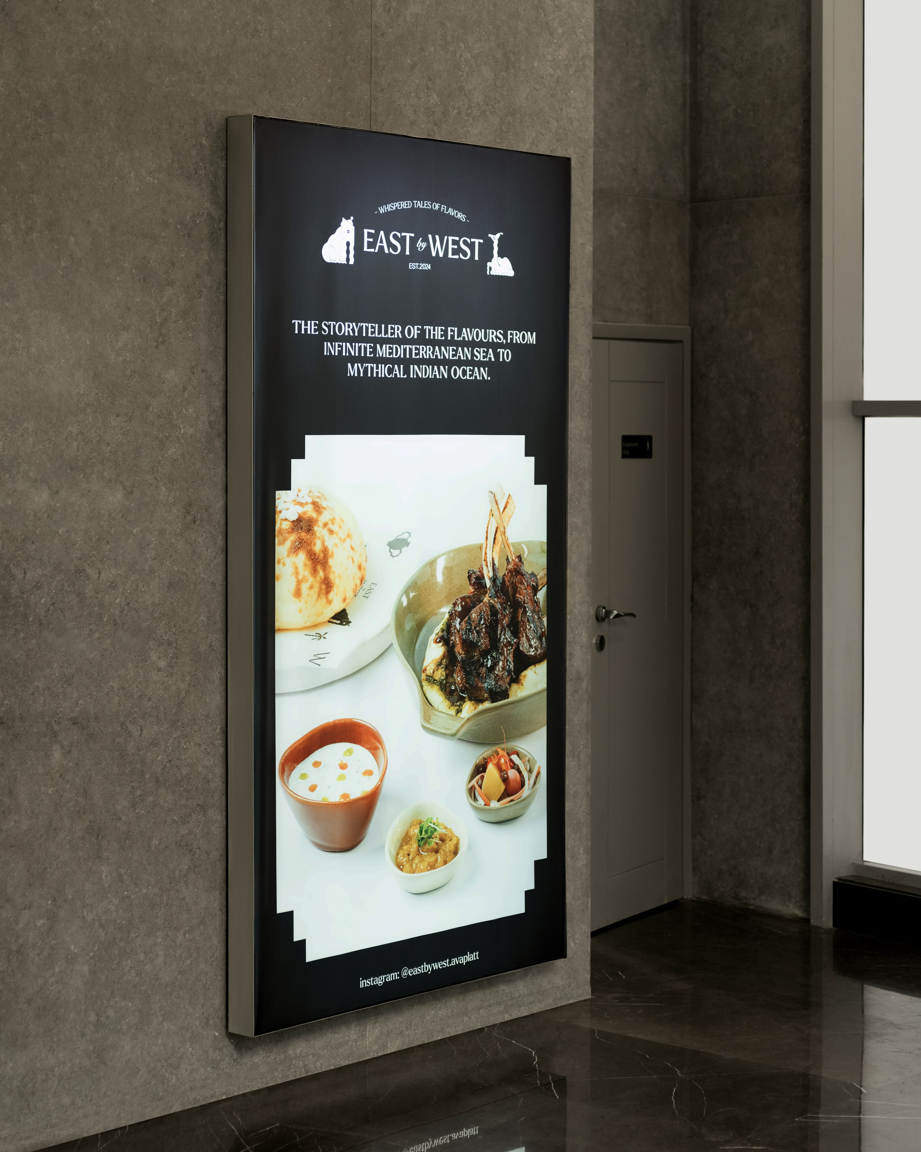 Illuminated East by West wall poster on textured gray surface: mythical Indian Ocean flavors tagline, logo, lamb dish in naan with chutney bowls beside restroom door.