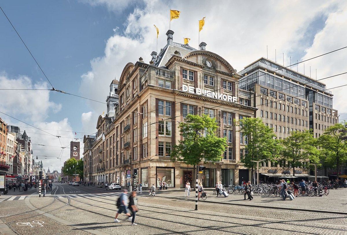 Uitzicht op de gevel van warenhuis De Bijenkorf in Amsterdam, met opvallende gele vlaggen en een historische architectuur. De foto toont drukke straatactiviteit met voetgangers, fietsers en winkelaars op de stoep, wat de levendige sfeer van de stad benadrukt. De Bijenkorf is een iconische bestemming voor luxe winkelen en biedt een breed scala aan producten. Deze afbeelding is ideaal voor het benadrukken van de centrale ligging en het winkelplezier in Amsterdam.