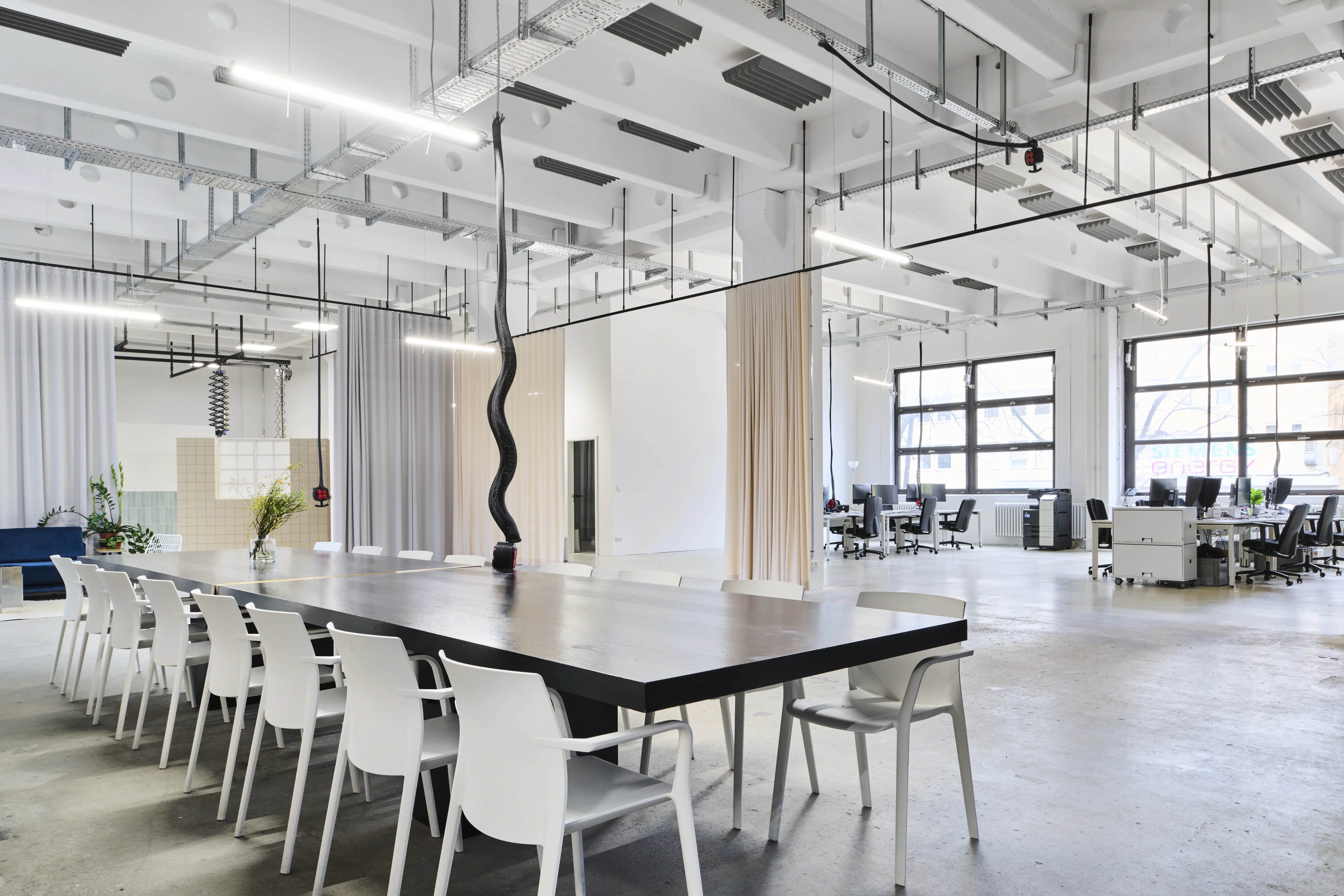 Open loft of the FLACONI CONTENT STUDIO with a long black meeting table and white chairs, curtain dividers in grey and beige and desk areas by the window front.