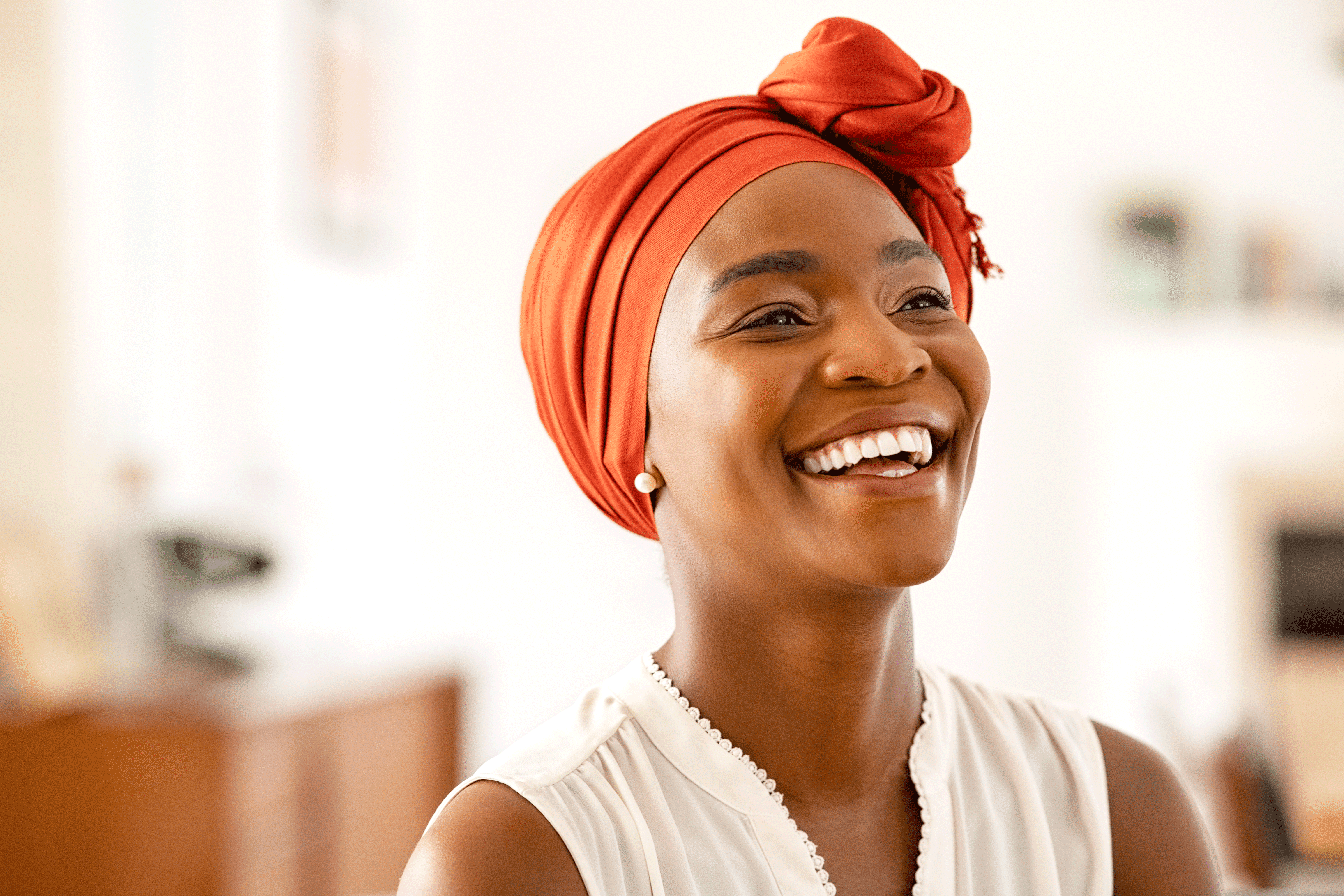 A woman with a bright, joyful smile looks off-camera while wearing a vibrant orange headwrap and a white sleeveless top. The background is a softly blurred interior, focusing attention on her expression and the detail of her headpiece.