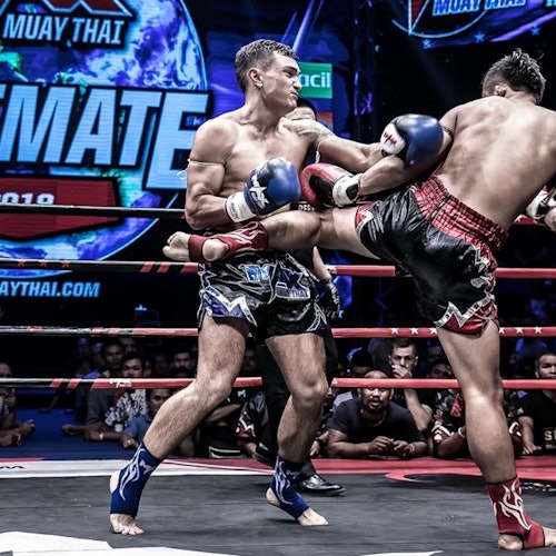 在擂台上，两名泰拳选手正在比赛，其中一名用高踢攻击对方的身体，背景中有观众和活动标志。