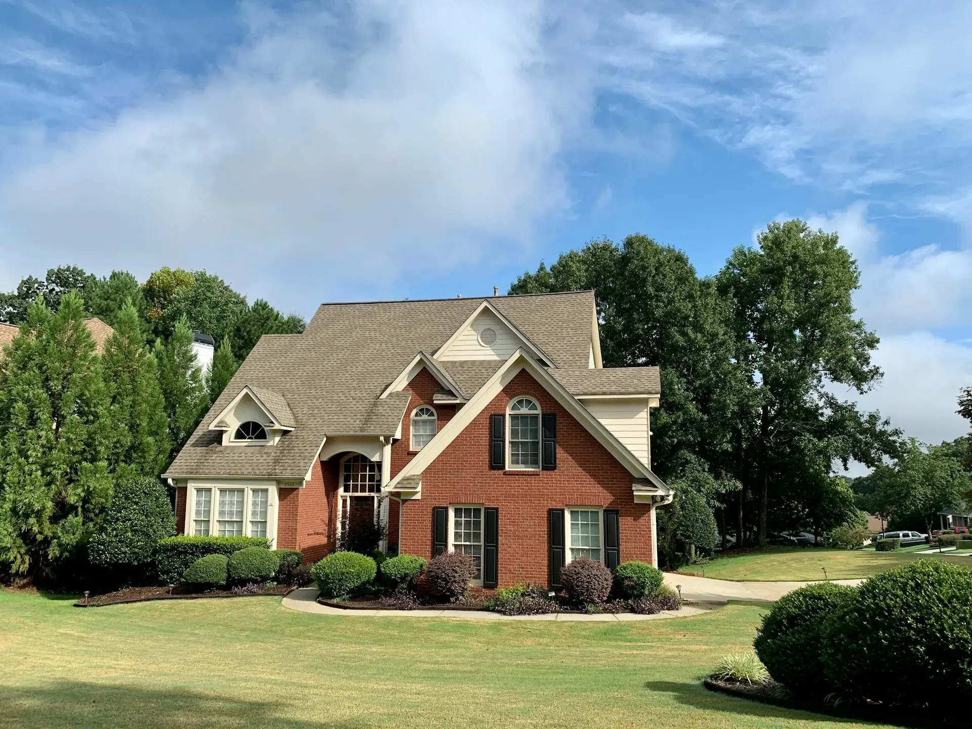 Red brick suburban house with a gabled roof, large windows, and a well-maintained front lawn surrounded by trees.