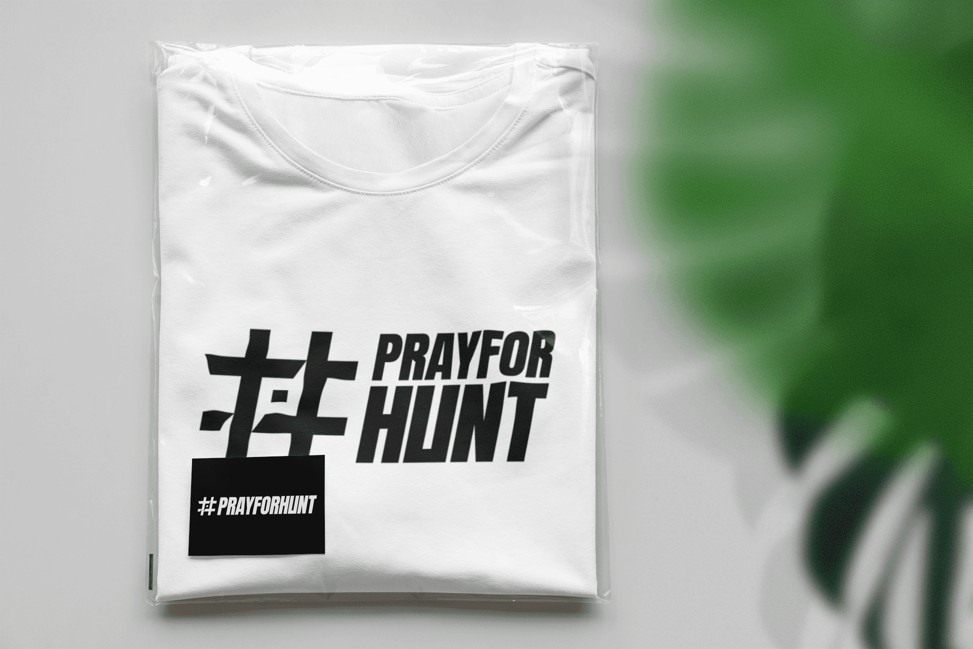 A white T-shirt neatly folded and packaged in a clear plastic wrap, displaying the '#PRAYFORHUNT' logo prominently in bold black print. The logo includes a stylized hashtag symbol and the words 'PRAY FOR HUNT' in an impactful font. A small black card placed on top of the T-shirt features the same logo, adding a cohesive branding element. The scene is set on a clean, light surface with a soft-focus green leaf partially visible in the corner, creating a fresh and inviting look.