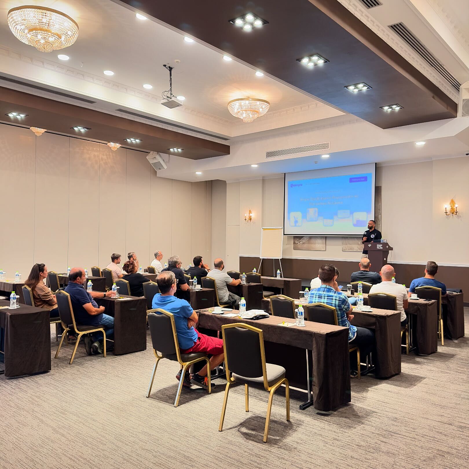 Two men sit at a wooden table with laptops, under a large red lamp. A woman walks by a window. The scene is calm, set against a red-brown wall.