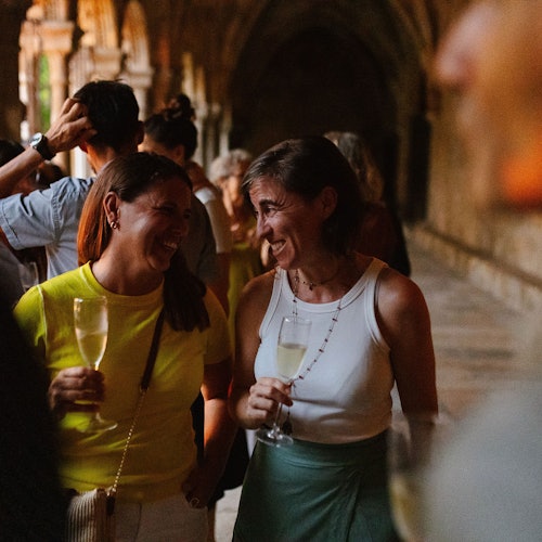 Two women holding glasses of champagne while conversing and smiling in a dimly lit, arched hallway with other people in the background.