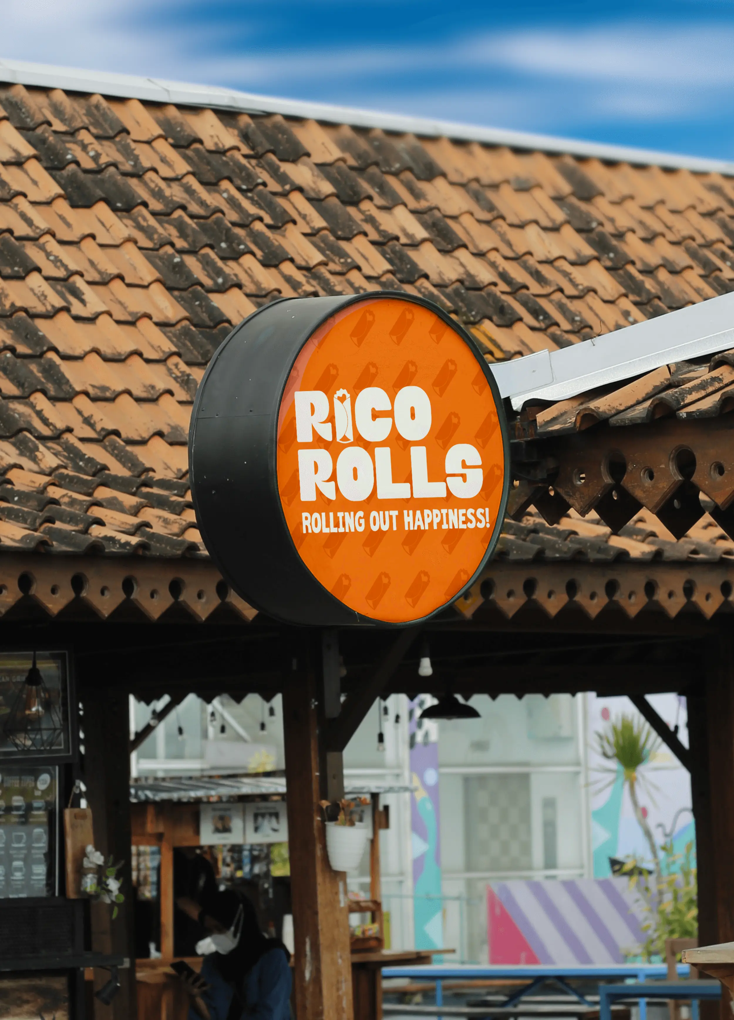 Rico Rolls circular storefront sign with the tagline ‘Rolling Out Happiness!’ displayed on an orange background, mounted on a rustic tiled-roof exterior.