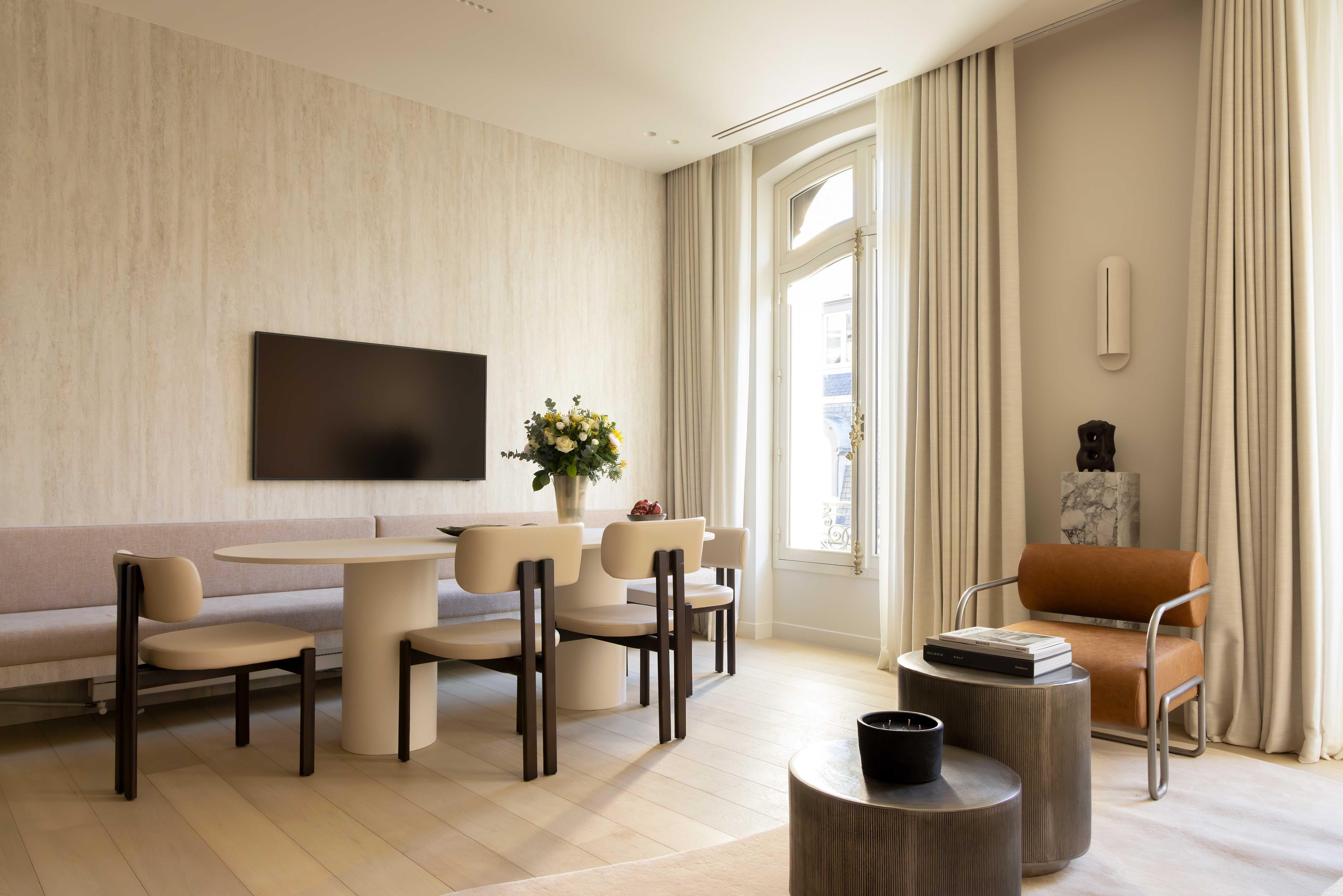 Espace salle a manger avec table ronde, chaises design beiges, banquette, television murale et mur texture sable