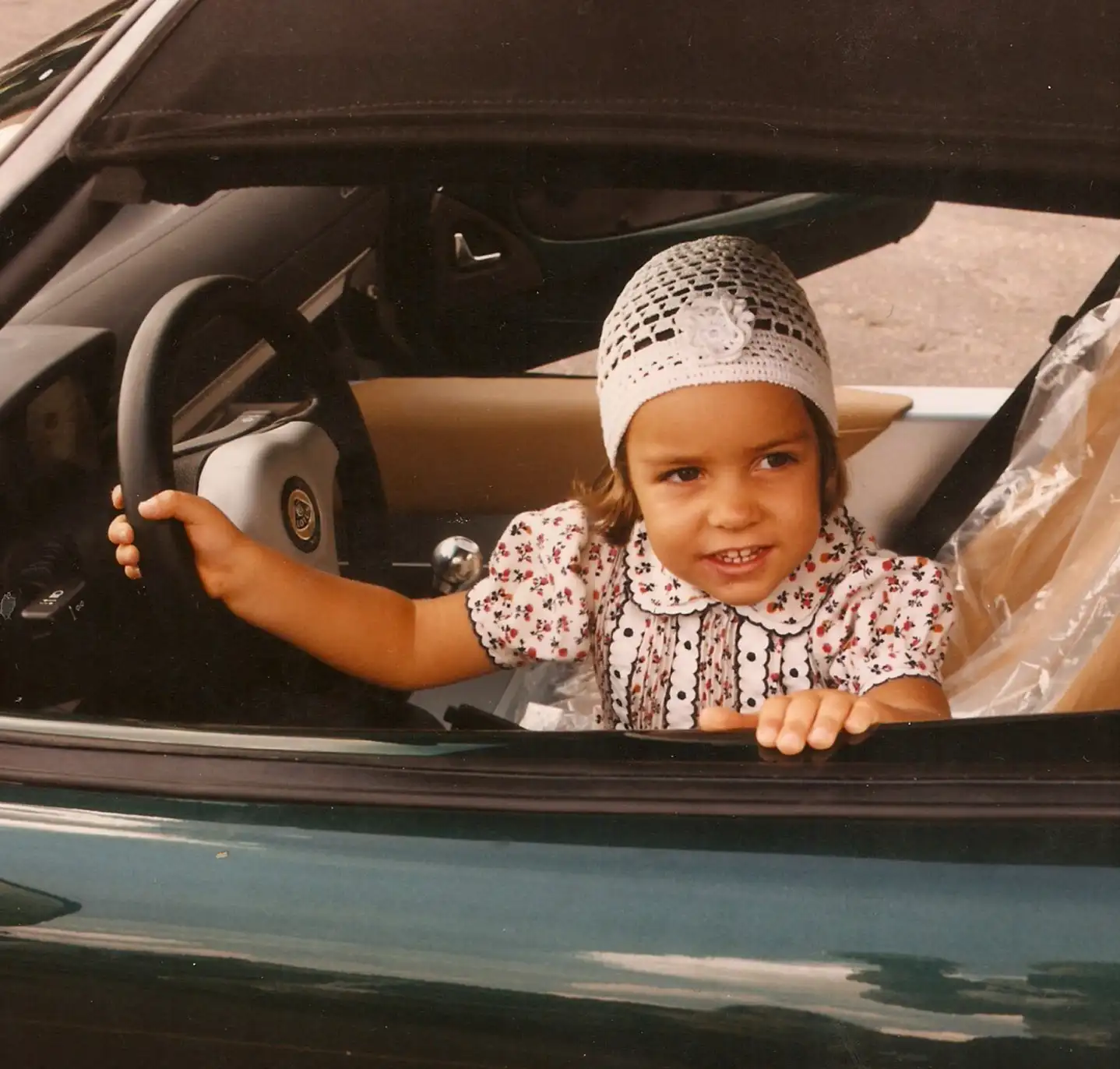 A young Elisa Artioli wearing a floral dress and a crocheted white headscarf sits in the driver’s seat of a green convertible Lotus sports car, smiling and holding the steering wheel.
