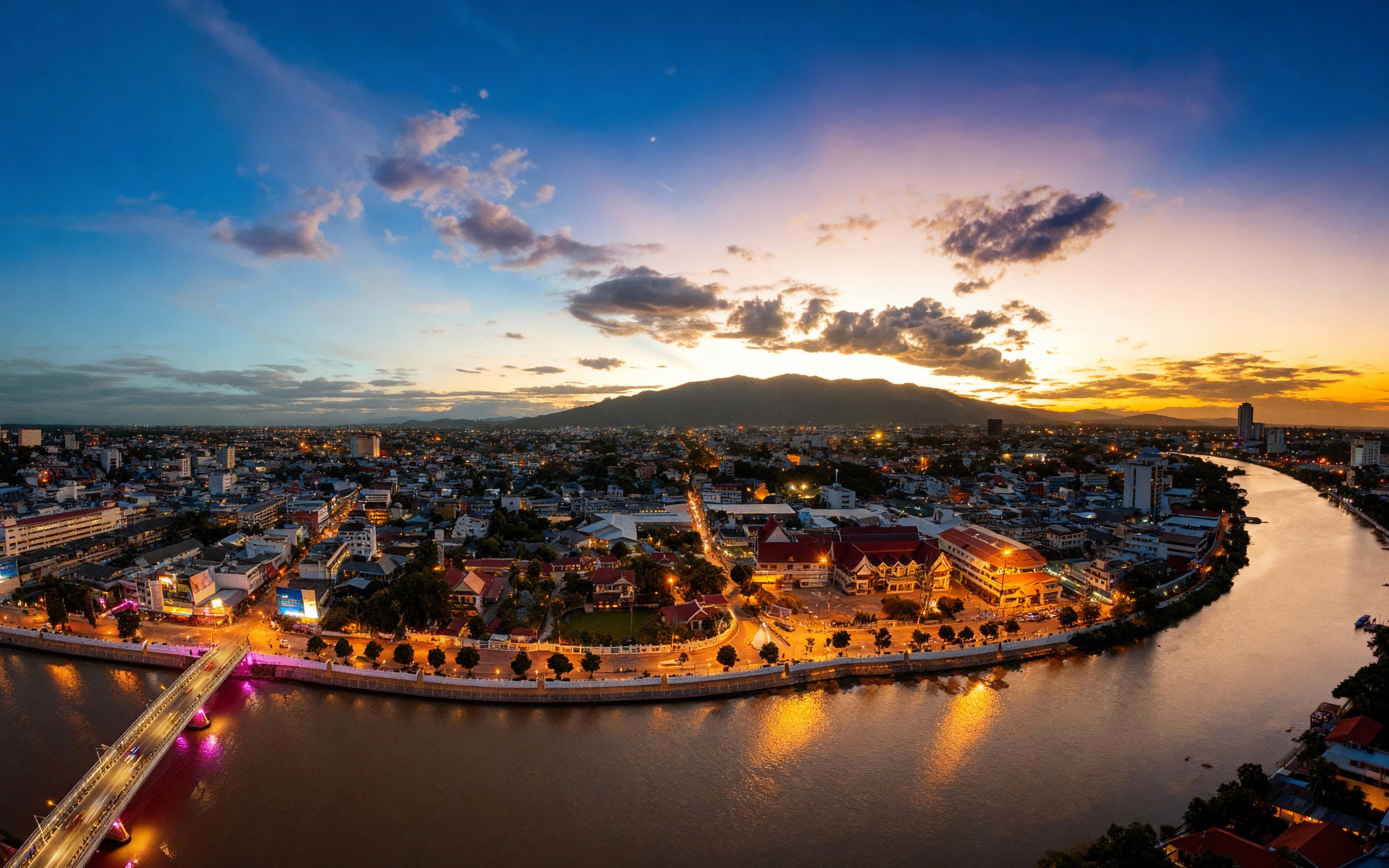 Aerial sunset view over Chiang Mai city with the Ping River curving through illuminated streets and Doi Suthep mountain silhouetted in the distance against a golden sky.
