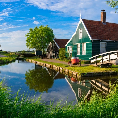 Green house with red roof by a calm canal, white footbridge, and lush greenery under a blue sky with scattered clouds.