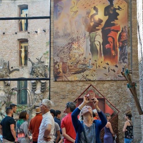 קהל של אנשים עומד מול בניין עם אמנות סוריאליסטית, בעוד שאדם אחד מצלם עם סמארטפון.